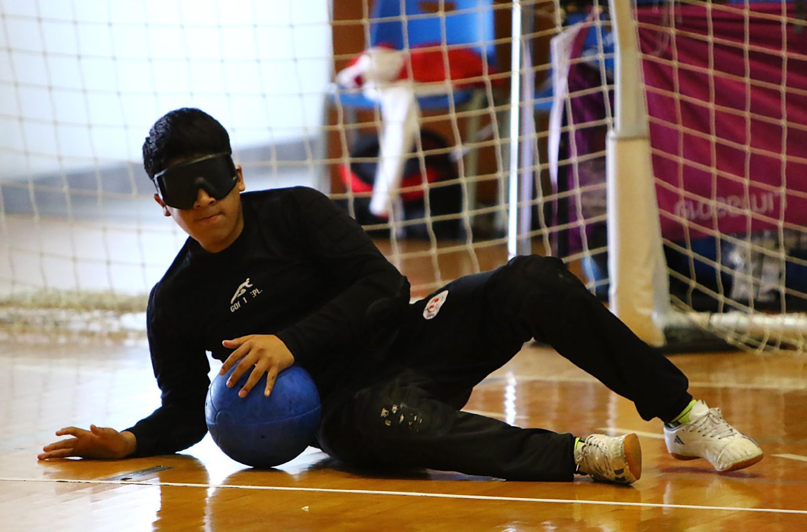 Jugadora peruana de goalball con antifaz, tendida en el piso junto al arco y abrazando el balón
